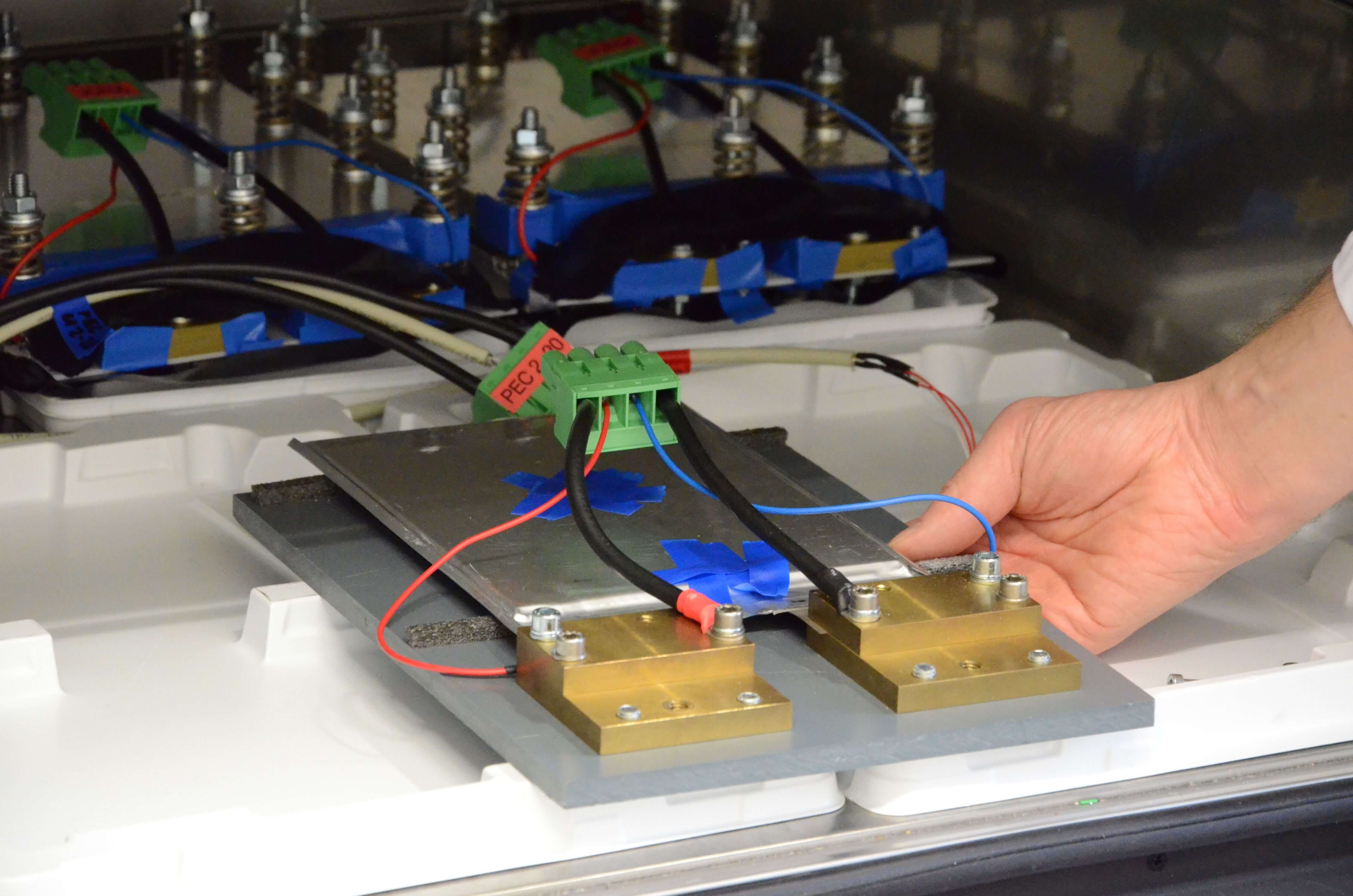 Eine Batteriezelle, die in einen Klimaschrank gelegt wird, wo sie an einen Tester angeschlossen und vermessen wird (im Hintergrund weitere, druckbelastete Batteriezellen) 